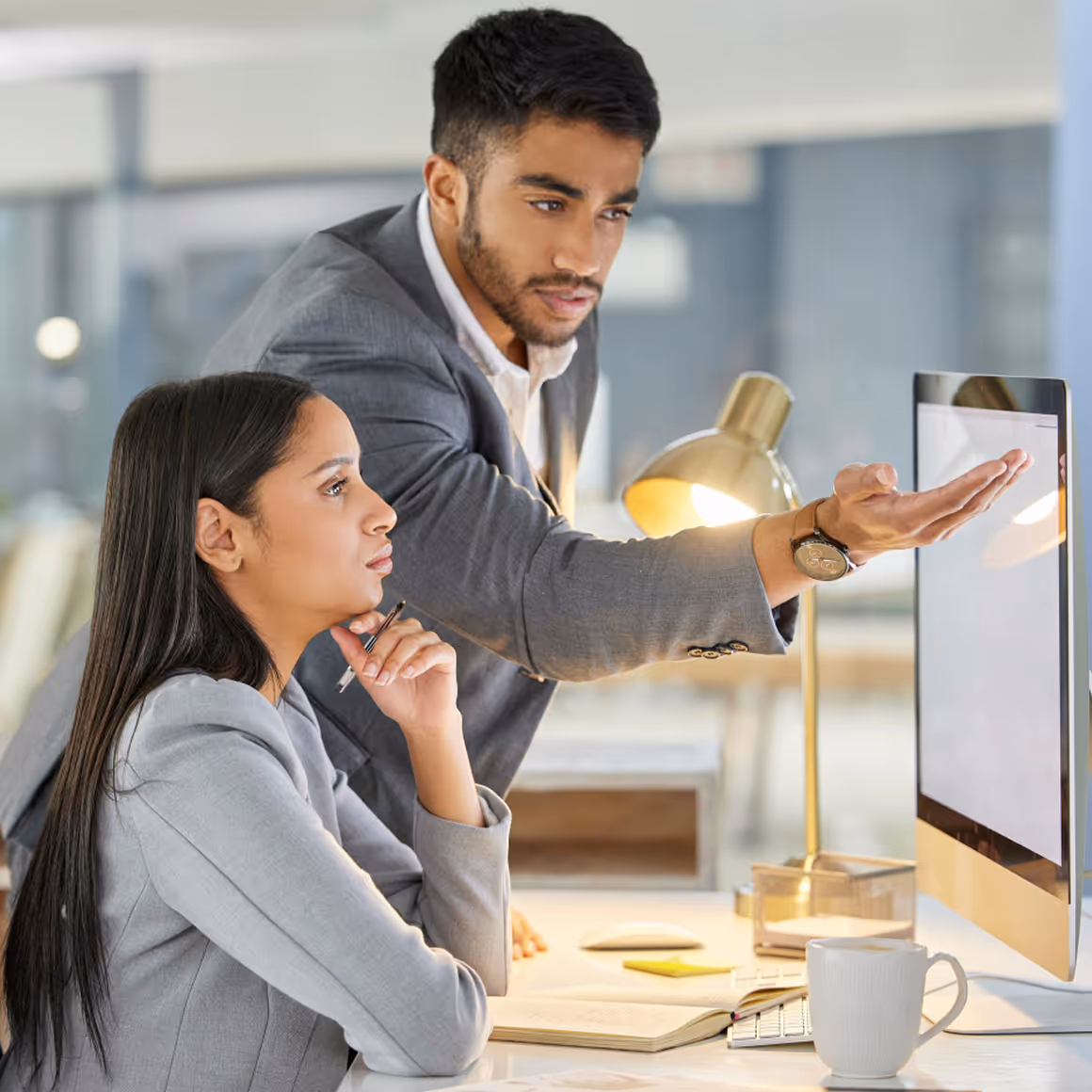 Two business colleagues in gray suits focus on a desktop computer screen in an office.