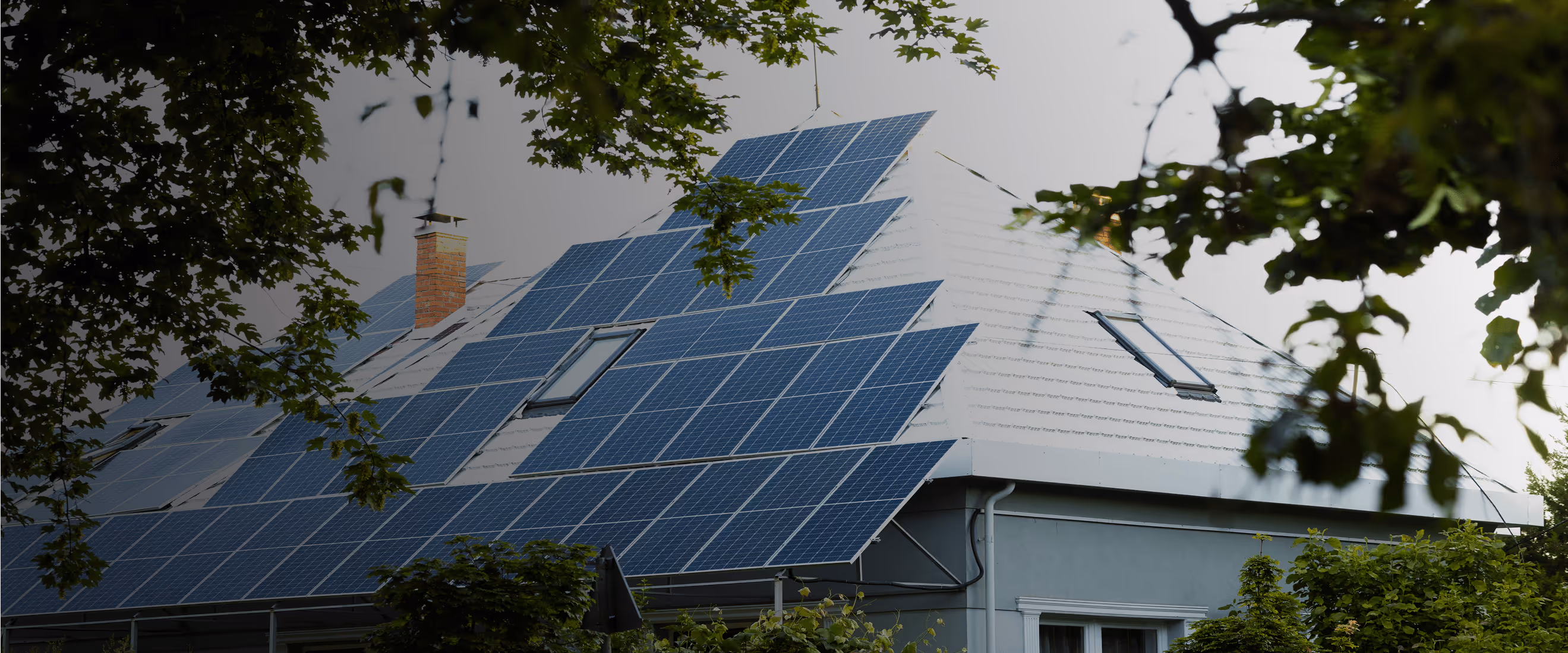House roof partially covered with blue solar panels and surrounded by green tree leaves.